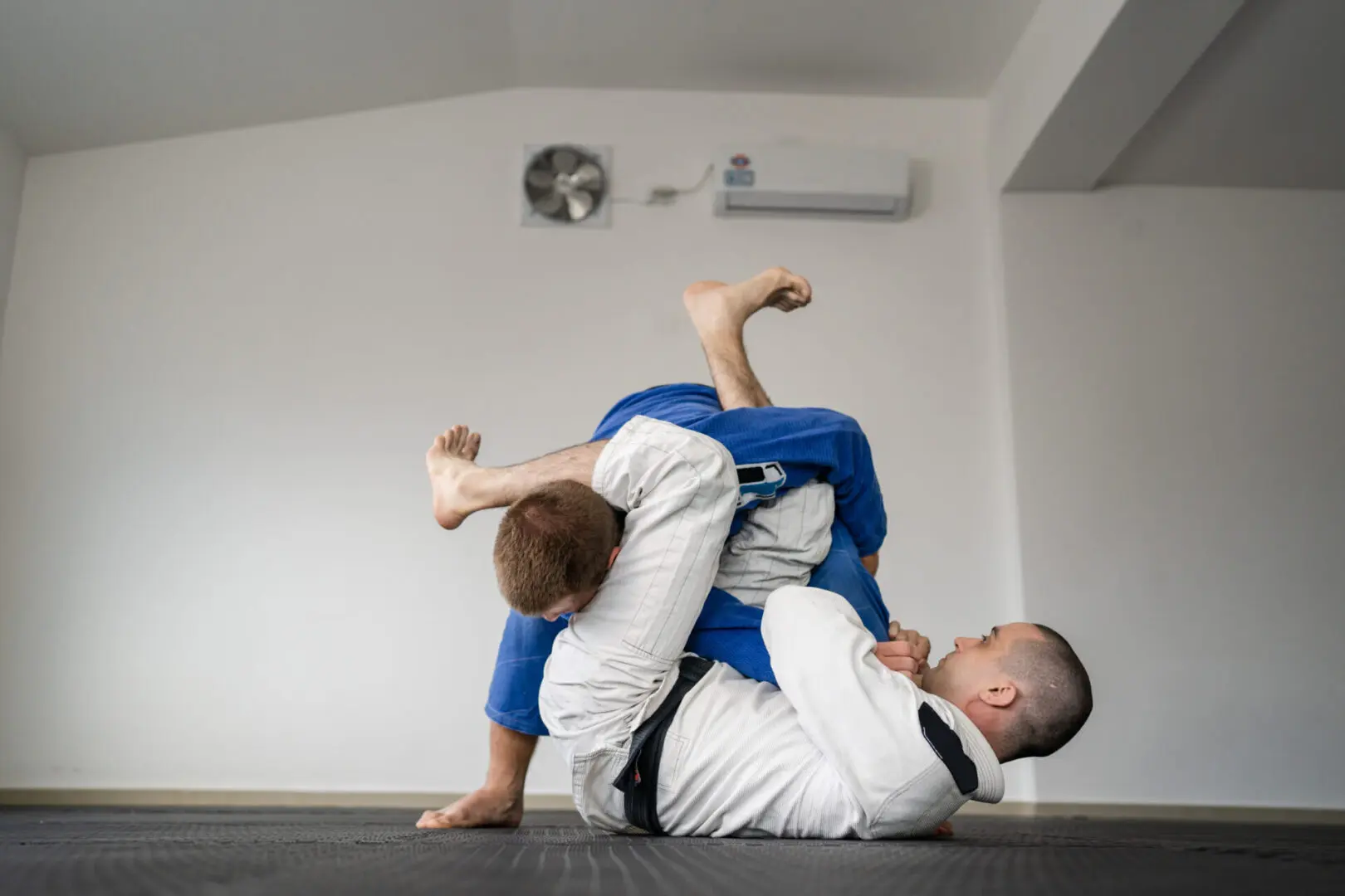 Two people practicing Brazilian jiu-jitsu on a mat, with one person wearing a blue gi executing a ground technique on a person in a white gi.