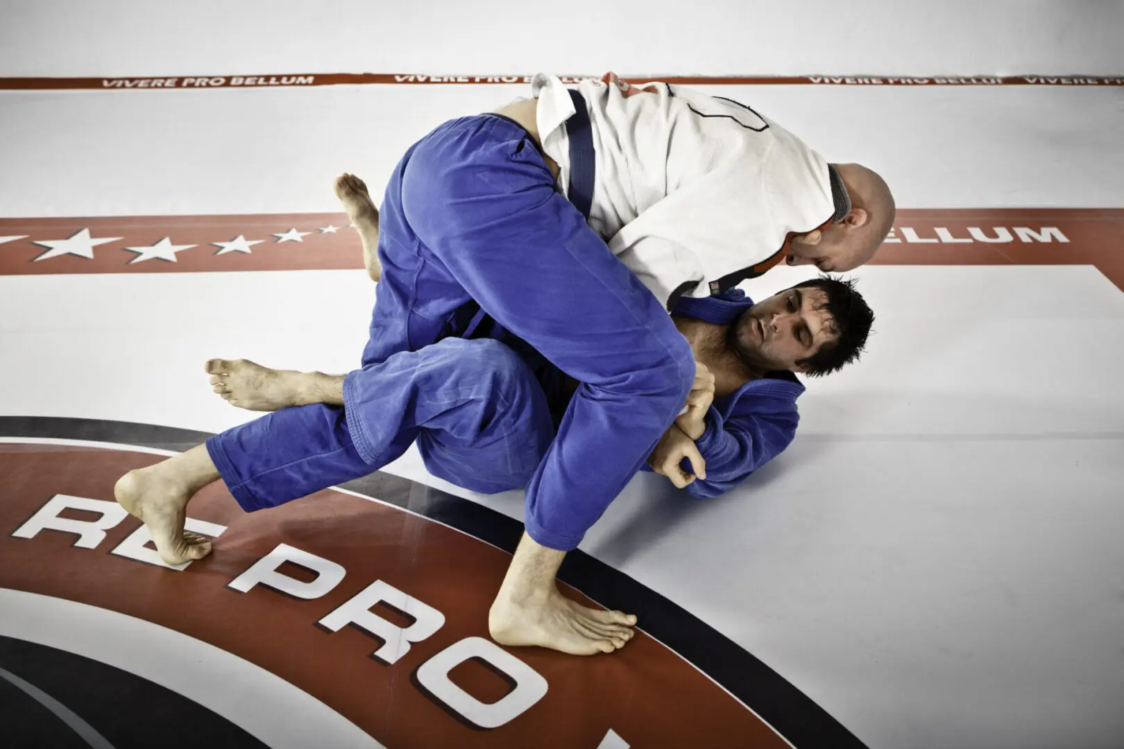 Two men in gi uniforms practice Brazilian Jiu-Jitsu on a mat. One is in a top position, while the other is on his back, defending. The mat has text and star designs.