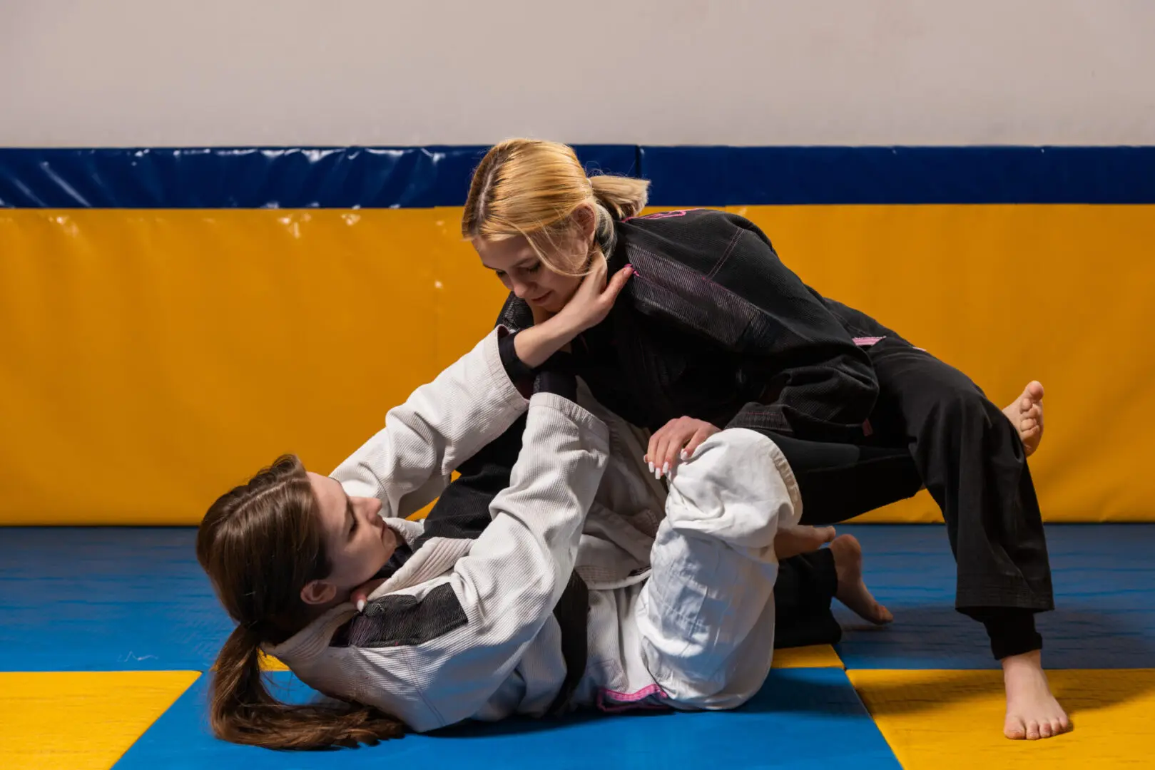 Two martial artists practicing on a mat. One, in a white gi, is on the ground, while the other, in a black gi, is leaning over to apply a technique.