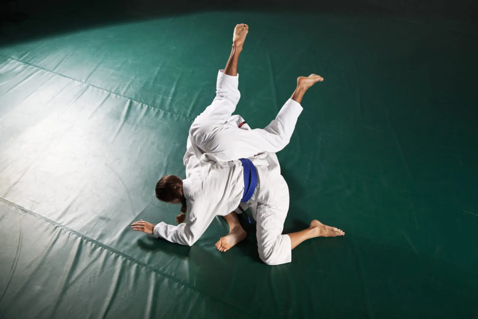 Two individuals in white martial arts uniforms practice grappling on a green mat, with one executing a throw.