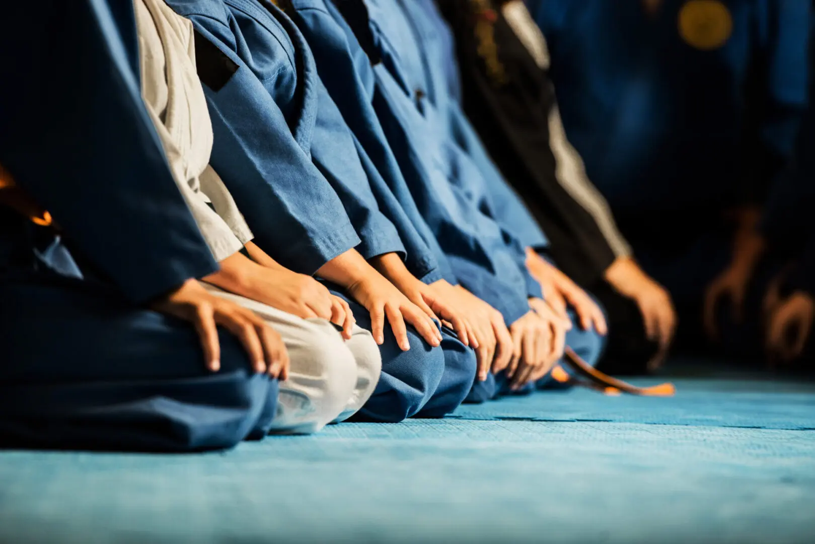 People in blue martial arts uniforms kneeling on a blue mat, with their hands resting on their knees.