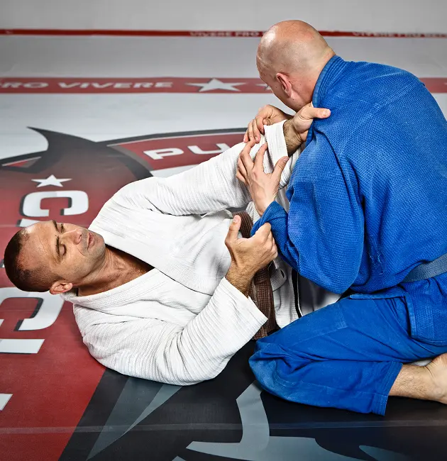 Two men in martial arts uniforms practicing jiu-jitsu on a training mat, one is on his side holding the other's arm, while the other kneels and stabilizes himself.
